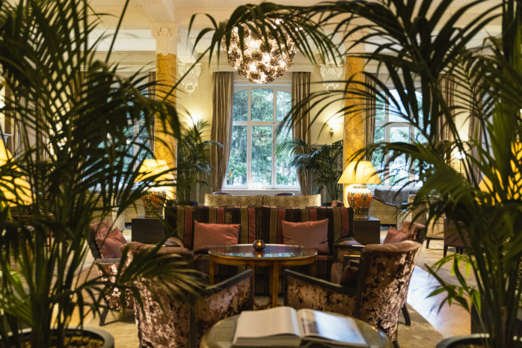 Stille Betrachtungen: Friedliche Zufluchten in historischen Hotels in Europa 7 Eine gemütliche Sitzecke mit Plüschsesseln, einem runden Tisch in der Mitte und einem großen Kronleuchter, umgeben von hohen Pflanzen und beleuchtet von warmem Licht aus Tischlampen, mit einem großen Fenster im Hintergrund - das erinnert an das elegante Ambiente im Cresta Palace Hotel Celerina Schweiz.