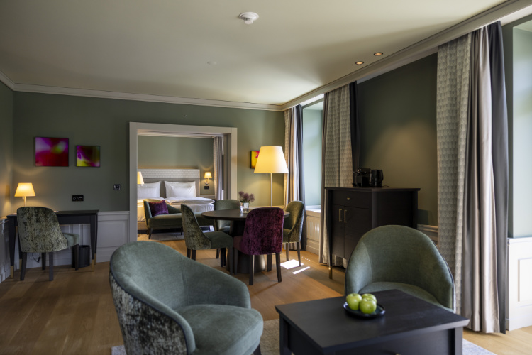 Une chambre d'hôtel bien éclairée du Cresta Palace Celerina comprend un coin salon avec deux chaises, une table ornée de pommes vertes, un bureau avec une lampe, et offre une vue sur la chambre à coucher avec son luxueux lit king-size, capturant l'essence des hôtels quatre étoiles en Suisse.