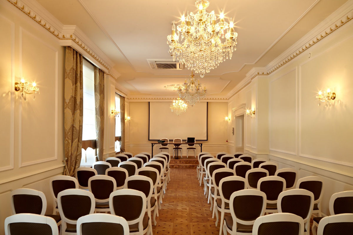 A conference room at Dwór Prezydencki with rows of chairs facing a presentation stage equipped with two chairs, a table, and a projector screen. The room has chandeliers, ornate wall sconces, and large windows with curtains.