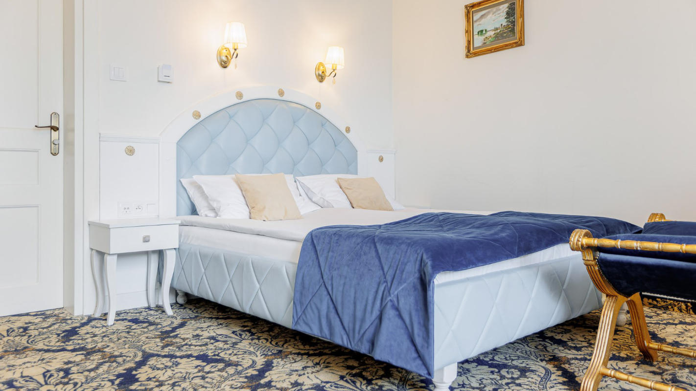 Image of a modern bedroom at Dwór Prezydencki, featuring a large bed with a tufted headboard, blue bedspread, and two pillows. The room boasts white walls, patterned carpet, wall-mounted lights, and a small nightstand.