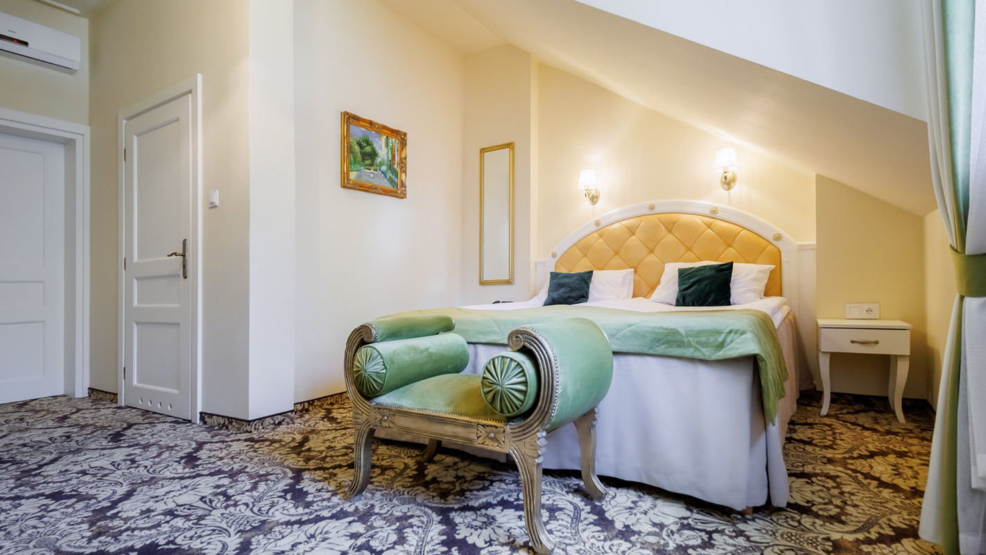 A neatly arranged hotel room at Dwór Prezydencki features a double bed, green and cream decor, two nightstands, a bench at the foot of the bed, a framed painting, and patterned carpet flooring.