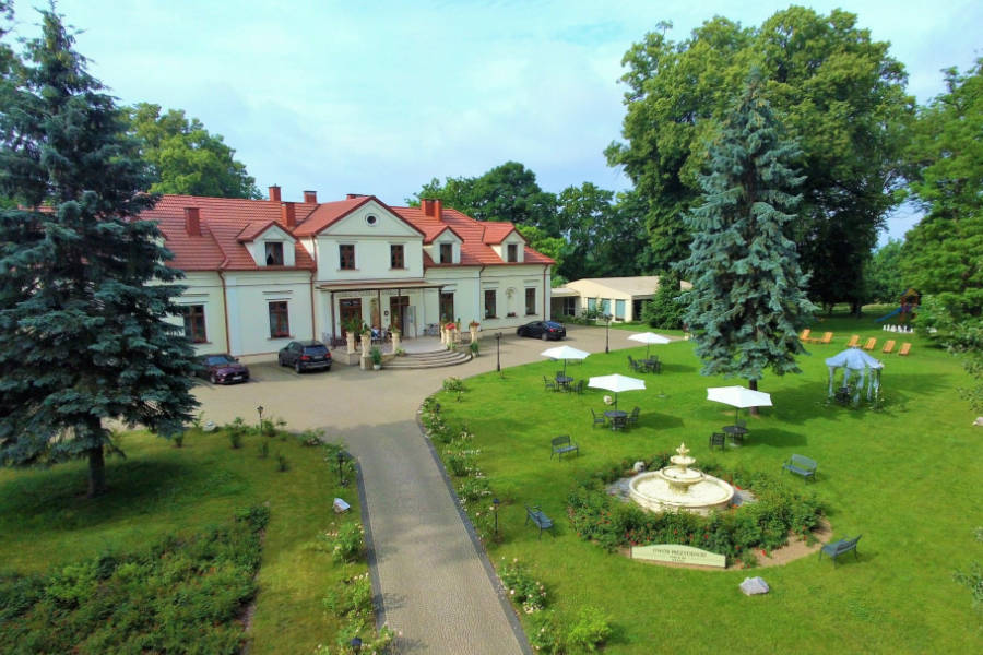 Aerial view of Dwór Prezydencki, a large white building with a red roof, surrounded by trees and a landscaped garden featuring a fountain, benches, and several white umbrellas.