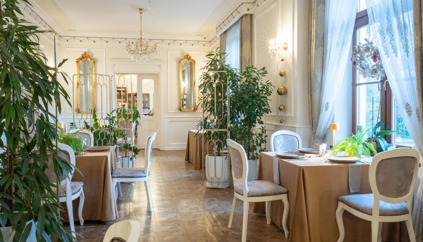 Elegant dining room at Dwór Prezydencki with white walls, chandeliers, mirrors, and large windows with sheer curtains. Tables are set with tan tablecloths, white chairs, and greenery decor. Several plants adorn the space.
