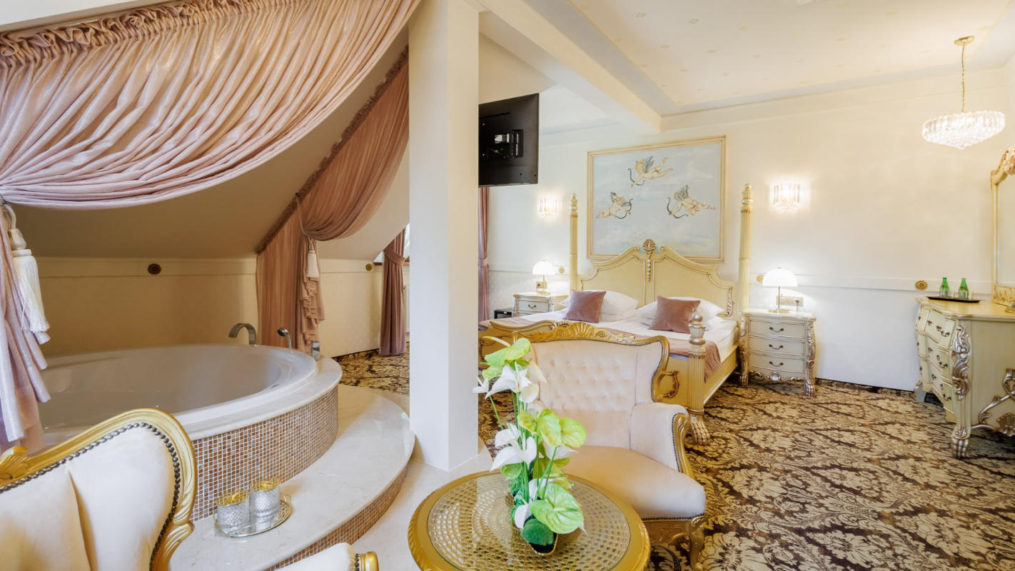 Elegant bedroom with ornate furniture, a large bed, a round bathtub, and gold-trimmed decor. The room at Dwór Prezydencki features a chandelier, floral carpet, and plush chairs near a small round table with a vase.