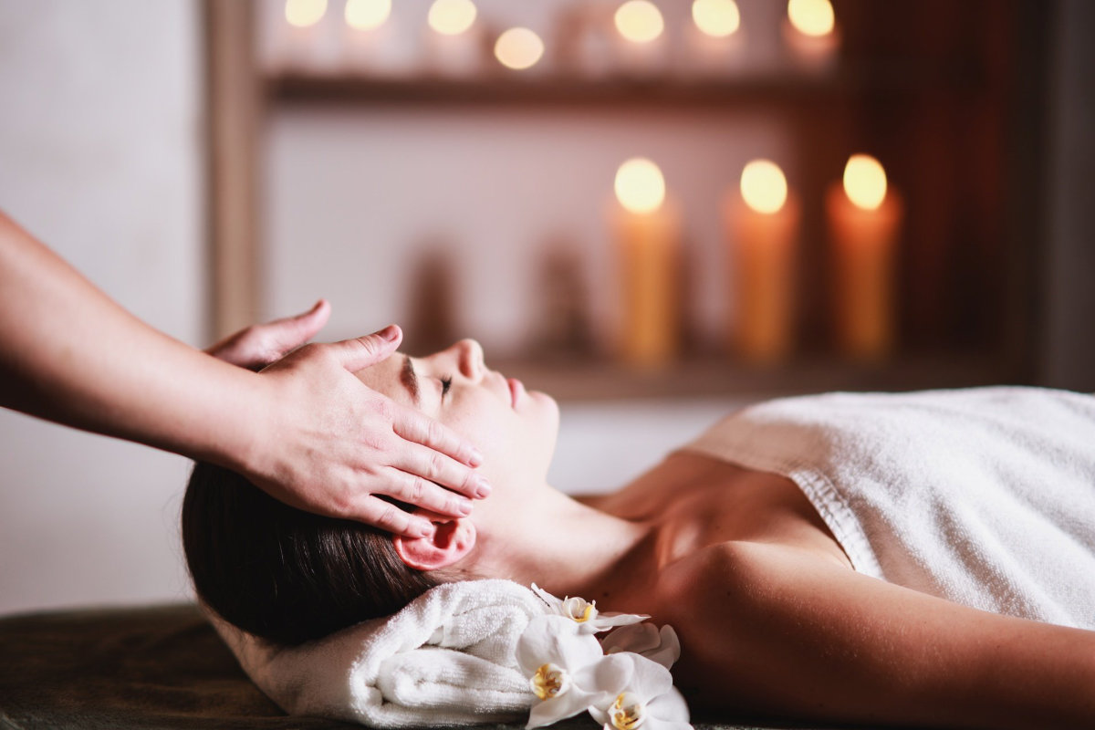 A person wrapped in a white towel lies on a massage table at Dwór Prezydencki, receiving a head massage. Candles are lit in the background.