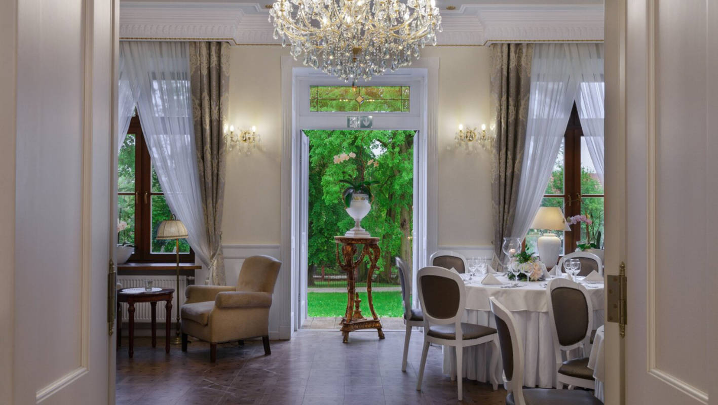Elegant dining room at Dwór Prezydencki with a chandelier, round dining table with chairs, armchairs, and large windows with sheer curtains opening to a garden view.