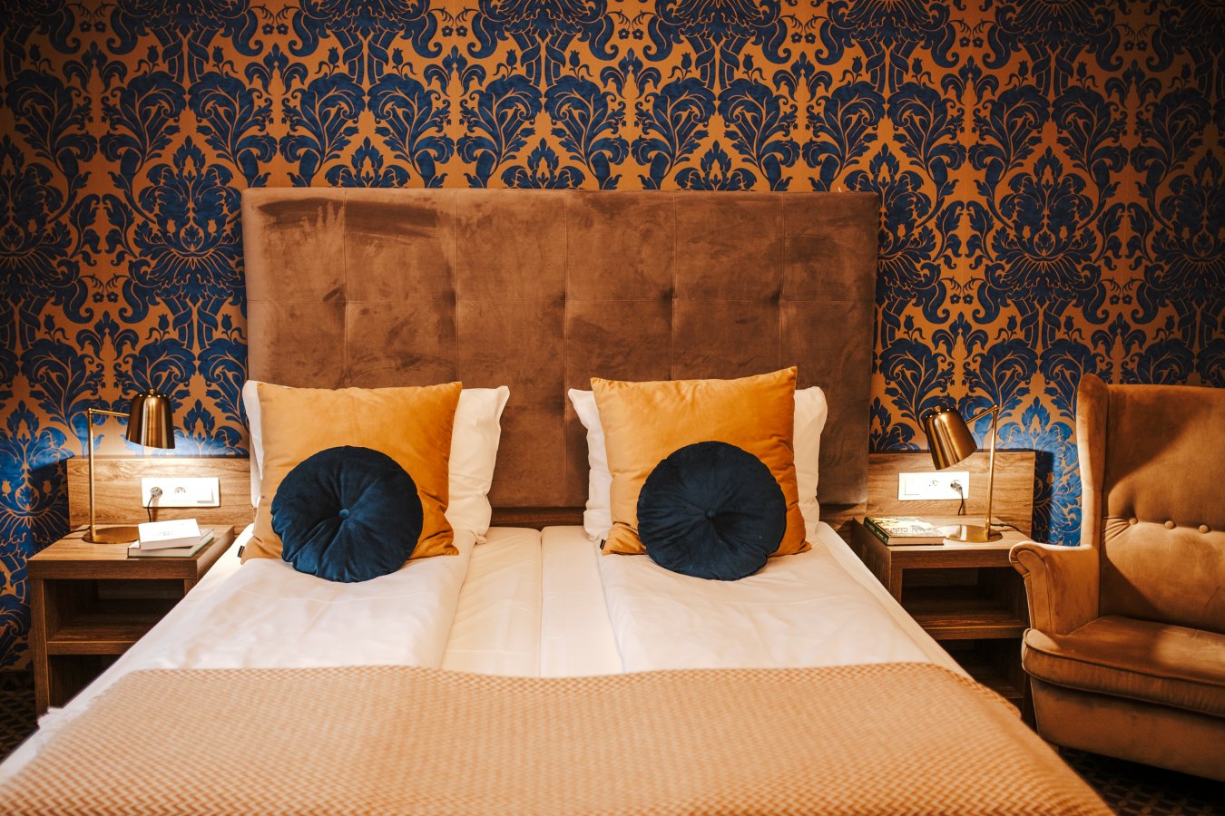 Ein Zimmer im Hotel Dębowy mit einem Doppelbett mit senfgelben und marineblauen Kissen, hölzernen Nachttischen mit Lampen und einem Sessel vor einer blau-gold gemusterten Wand.