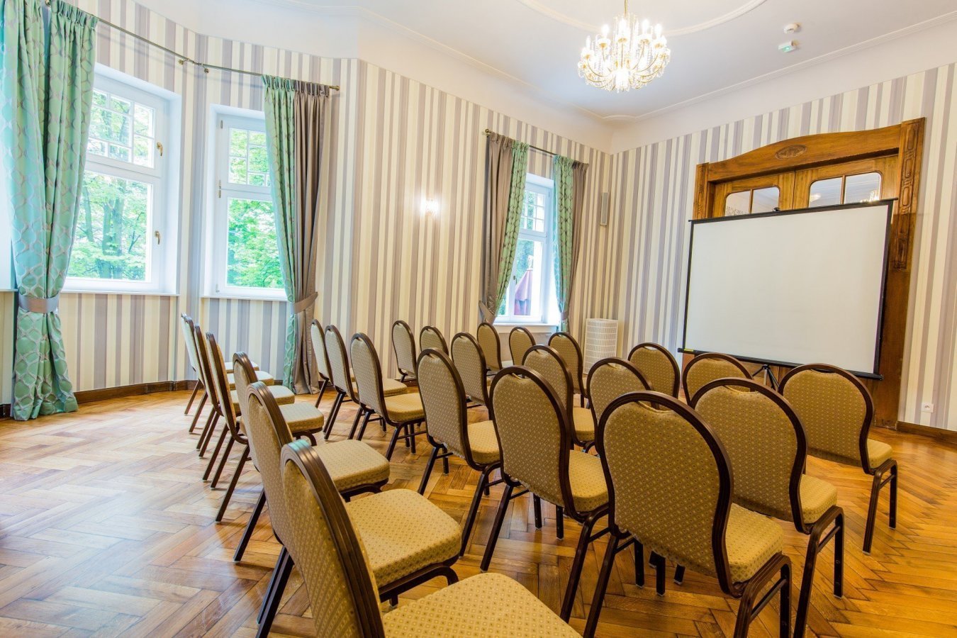Ein kleiner Konferenzraum im Hotel Dębowy ist mit beigen Stuhlreihen vor einer Projektionsfläche, gestreifter Tapete, Holzfußboden und mehreren Fenstern mit grünen Vorhängen ausgestattet.