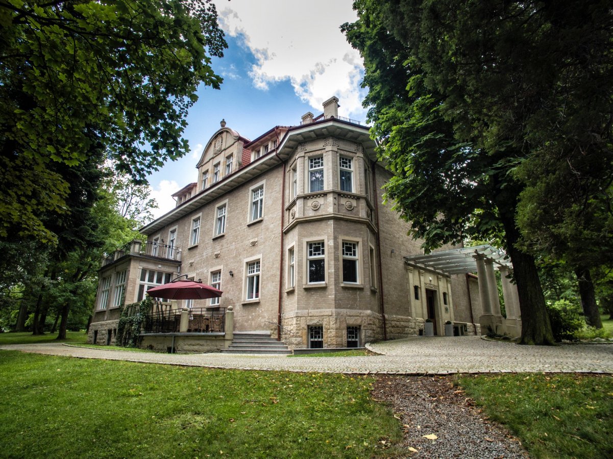 A large, historic brick mansion with a red roof sits surrounded by lush greenery and trees, featuring a patio with an umbrella and a gravel pathway—welcome to Hotel Dębowy.