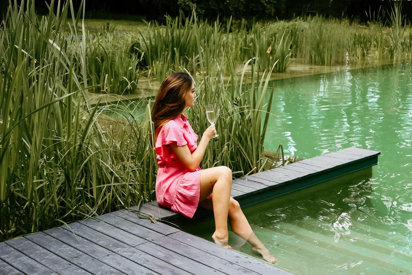 Eine Frau in einem rosafarbenen Kleid sitzt mit einem Glas in der Hand auf einem hölzernen Steg, der das Hotel Dębowy mit seinem hohen Gras umgibt, und hält die Füße ins Wasser.