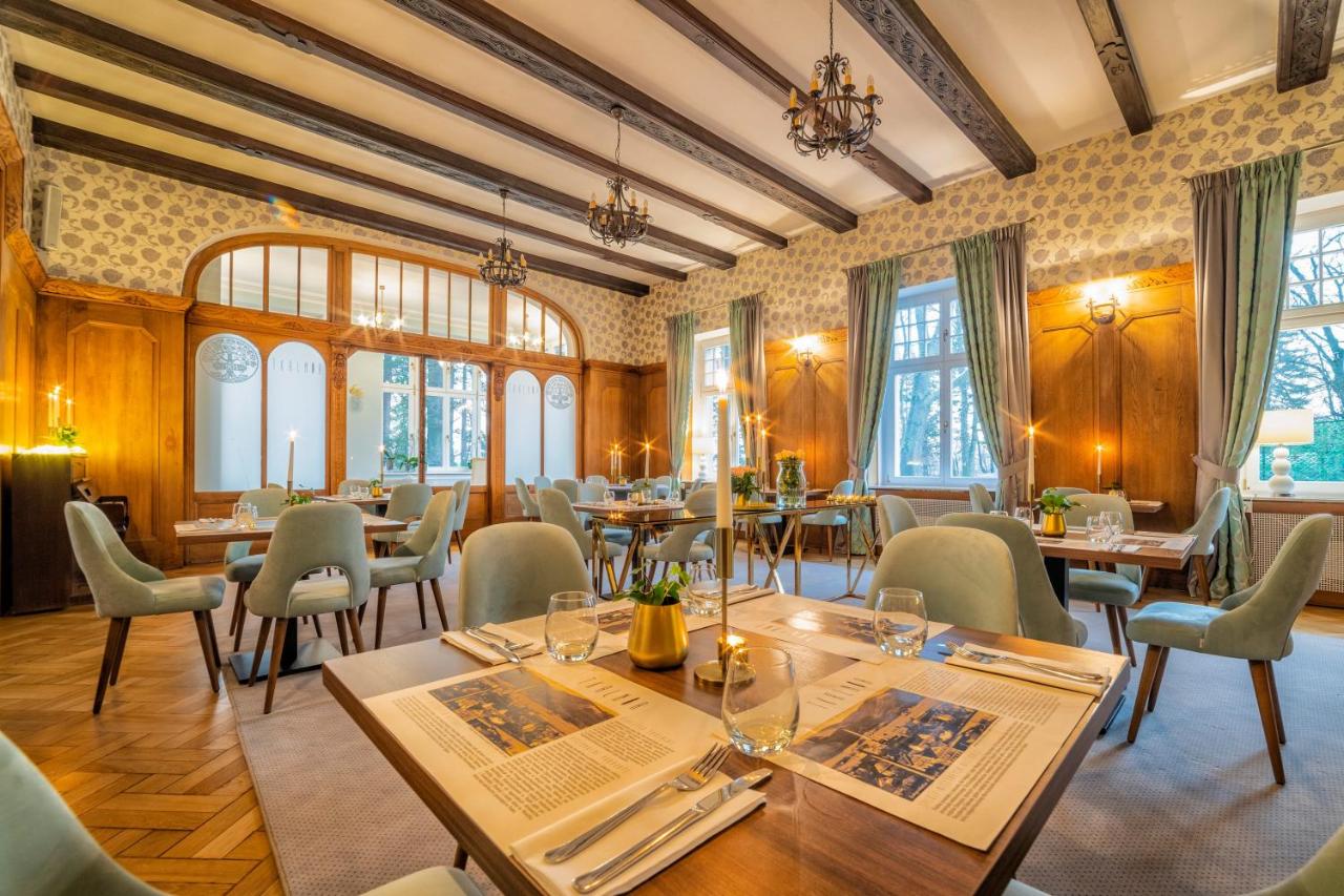 Ein elegant eingerichteter Speisesaal im Hotel Dębowy mit Holzwänden, gemusterten Tapeten und Kronleuchtern. Die Tische sind mit Kerzen, Besteck, Weingläsern und Tischsets im Zeitungsstil gedeckt. Das Licht fällt durch große Fenster.