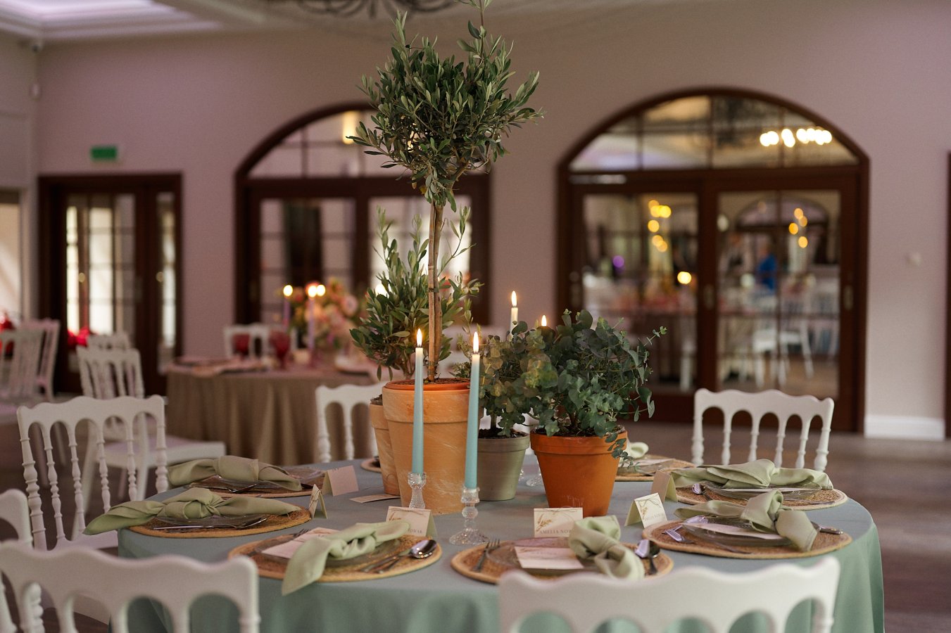 Gedeckter Empfangstisch für eine Veranstaltung im Hotel Dębowy, geschmückt mit Topfpflanzen, Kerzen und Platzdeckchen mit grünen Servietten. Im Hintergrund ein weiterer gedeckter Tisch und große Fenster mit Bogenrahmen, die der Atmosphäre Eleganz verleihen.