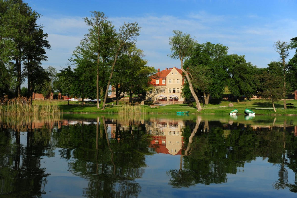 Una gran casa con paredes de color amarillo pálido y rojo, reminiscencia de la encantadora arquitectura de Olandia, se refleja en un tranquilo lago rodeado de altos árboles y barcos en un día soleado.