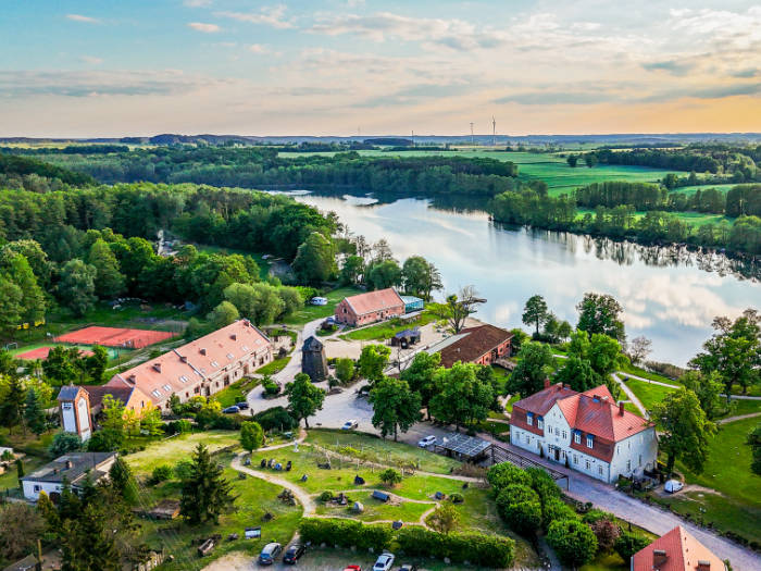 Luftaufnahme des ruhigen Seedorfs Olandia mit seinen roten Dächern, üppigen grünen Bäumen und einem klaren See, umgeben von einer weiten Landschaft unter einem strahlenden Himmel.