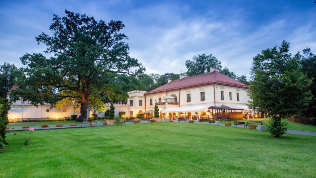 Europe’s most Historic Hotels 6 A large, two-story building with a red roof and white exterior stands next to a sprawling green lawn and tall tree, reminiscent of the charm found at Zamek Dubiecko, set against a cloudy sky.