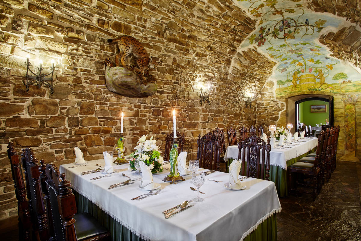 Un comedor con paredes de piedra, una larga mesa blanca con mantel preparada para comer, sillas de madera adornadas, apliques de pared, velas y un fresco en el techo que representa un paisaje que recuerda al Zamek Dubiecko. En la pared hay una gran estatua de un gato.