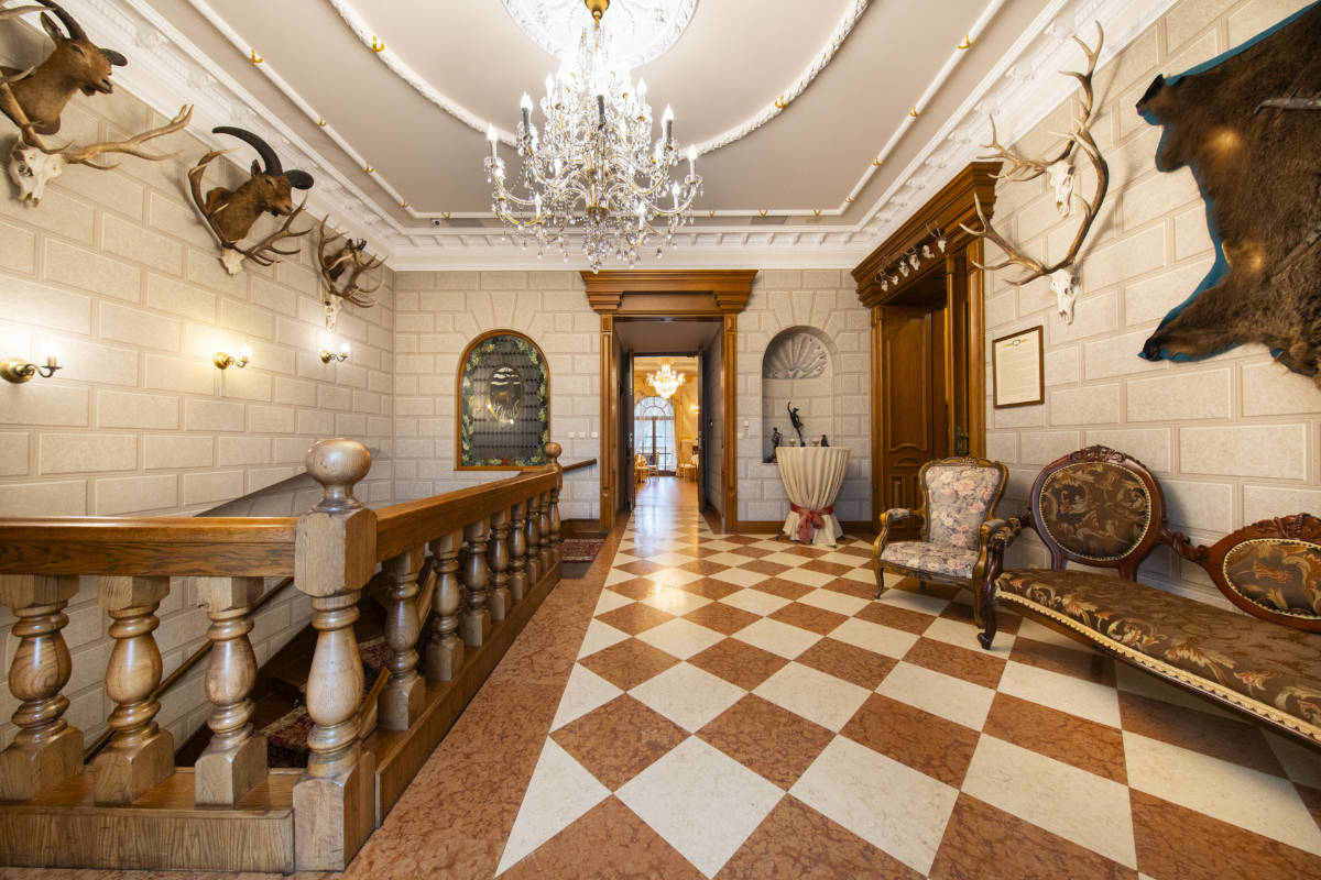 Un pasillo ornamentado con suelo a cuadros, lámparas de araña, cabezas de animales montadas en las paredes, muebles antiguos y una barandilla de madera que recuerda al Zamek Dubiecko.