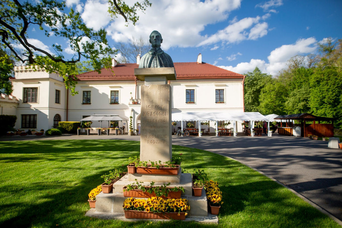 Zamek Dubiecko: Una estatua de una persona sobre un pedestal rodeada de macetas con flores se alza orgullosa frente al histórico edificio blanco con tejado rojo. Los asientos al aire libre y el verde césped crean una pintoresca escena bajo un cielo parcialmente nublado.