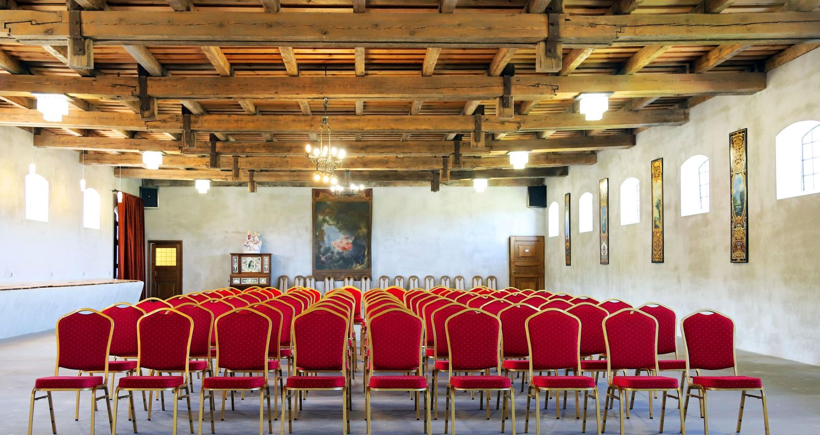 Ein geräumiger Saal im Kadyny Folwark Hotel mit hölzernen Deckenbalken, in dem Reihen von roten, gepolsterten Stühlen für eine Veranstaltung aufgestellt sind. Der Raum hat weiße Wände, mehrere Fenster und ein großes Gemälde an der Stirnseite.
