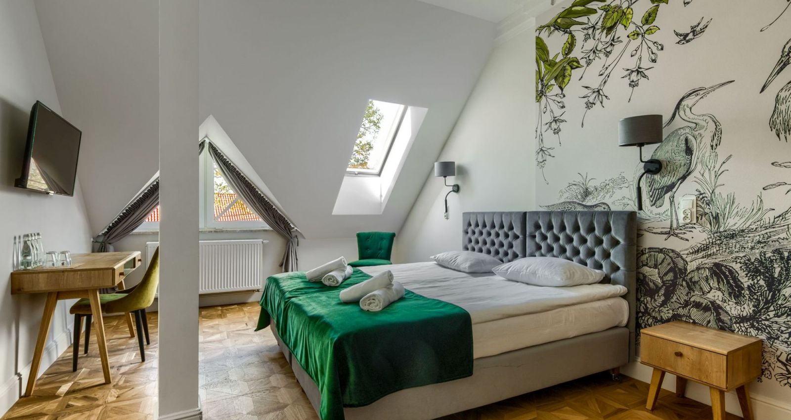 Ein modernes, vom Kadyny Folwark Hotel inspiriertes Schlafzimmer mit einem Doppelbett mit grüner Bettwäsche, einem an der Wand montierten Fernseher, zwei Oberlichtern, Holzfußboden, einem Schreibtisch und einem Wandgemälde mit Naturmotiven an der Wand.