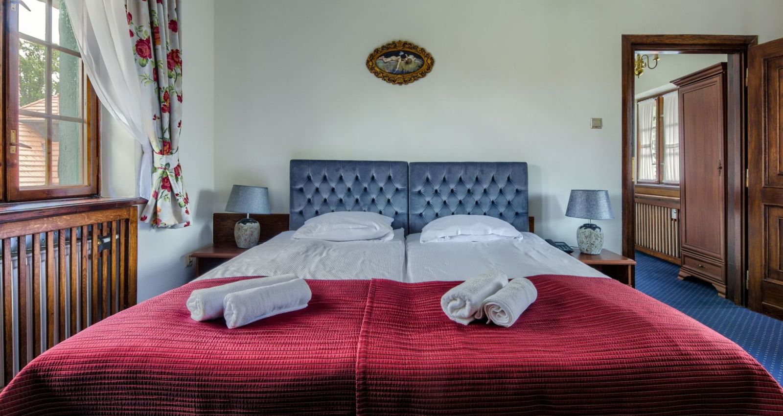 Ein ordentlich gemachtes Doppelbett mit einer roten Steppdecke, zwei gerollten Handtüchern und zwei passenden Nachttischlampen in einem gemütlichen Schlafzimmer mit geblümten Vorhängen, das an die charmante Einrichtung des Kadyny Folwark Hotels erinnert.