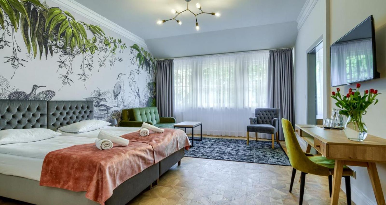 Ein modernes Zimmer im Kadyny Folwark Hotel verfügt über zwei Einzelbetten, ein Wandgemälde, zwei Stühle, einen Schreibtisch mit einer Vase mit roten Tulpen und einen Fernseher. Ein Fenster mit weißen Vorhängen bietet einen Blick auf die grüne Landschaft im Hintergrund.