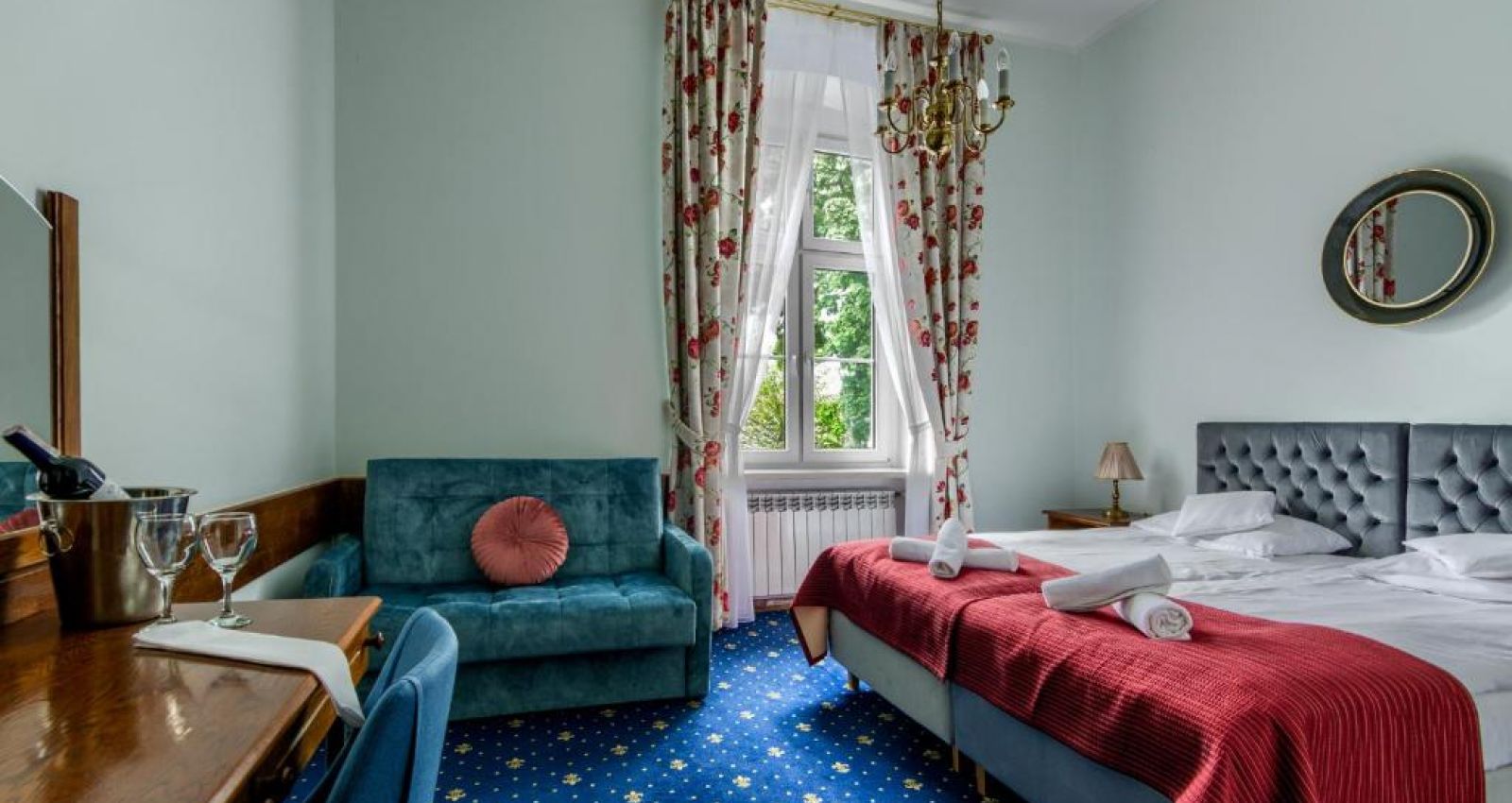 Ein Zimmer im Kadyny Folwark Hotel mit einem Doppelbett mit roten Decken, einem tealfarbenen Sofa, einem hölzernen Schreibtisch mit einem Spiegel, einer Flasche Wein mit zwei Gläsern und geblümten Vorhängen.