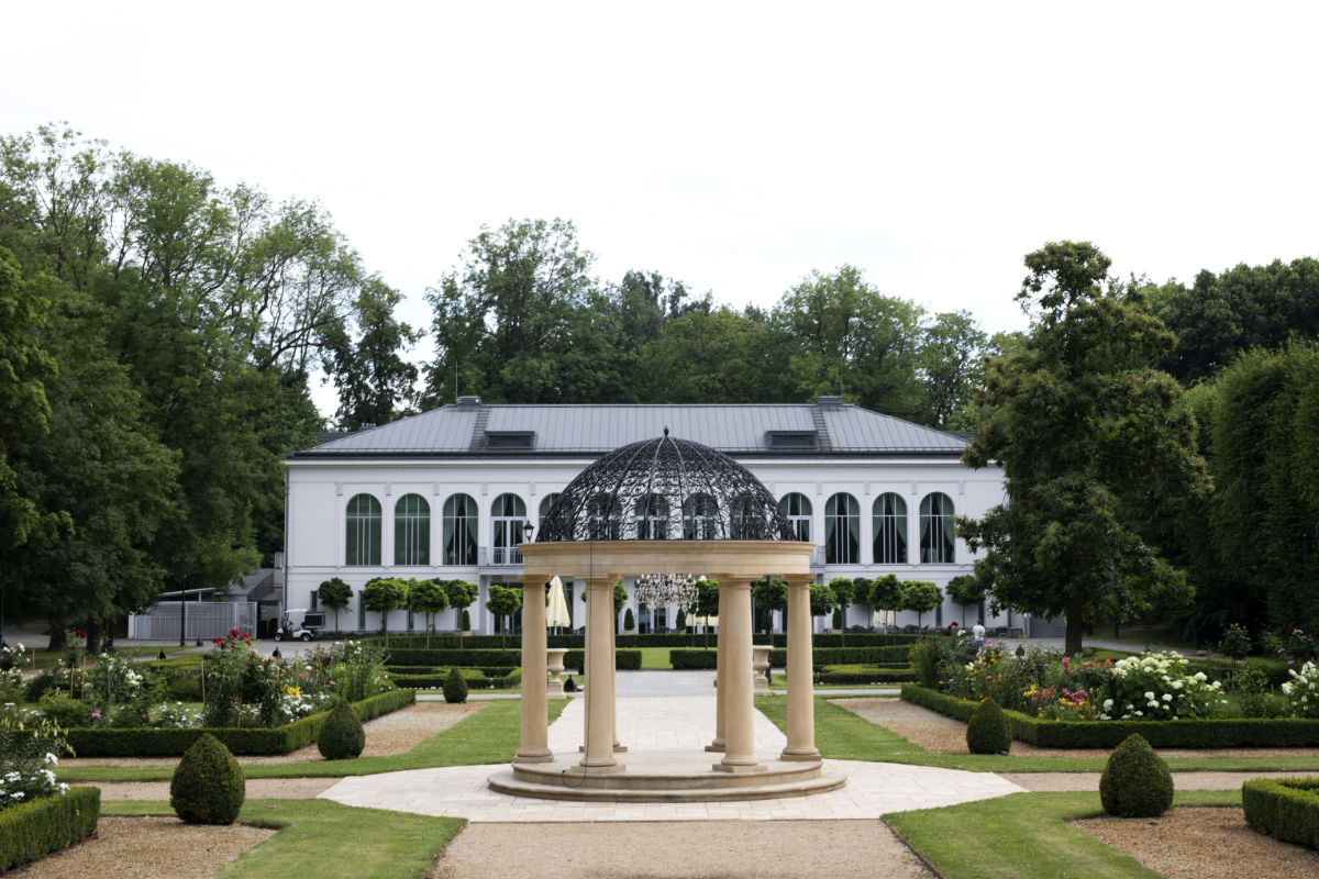 Un giardino paesaggistico con un gazebo bianco in primo piano e un grande edificio bianco con finestre ad arco sullo sfondo.