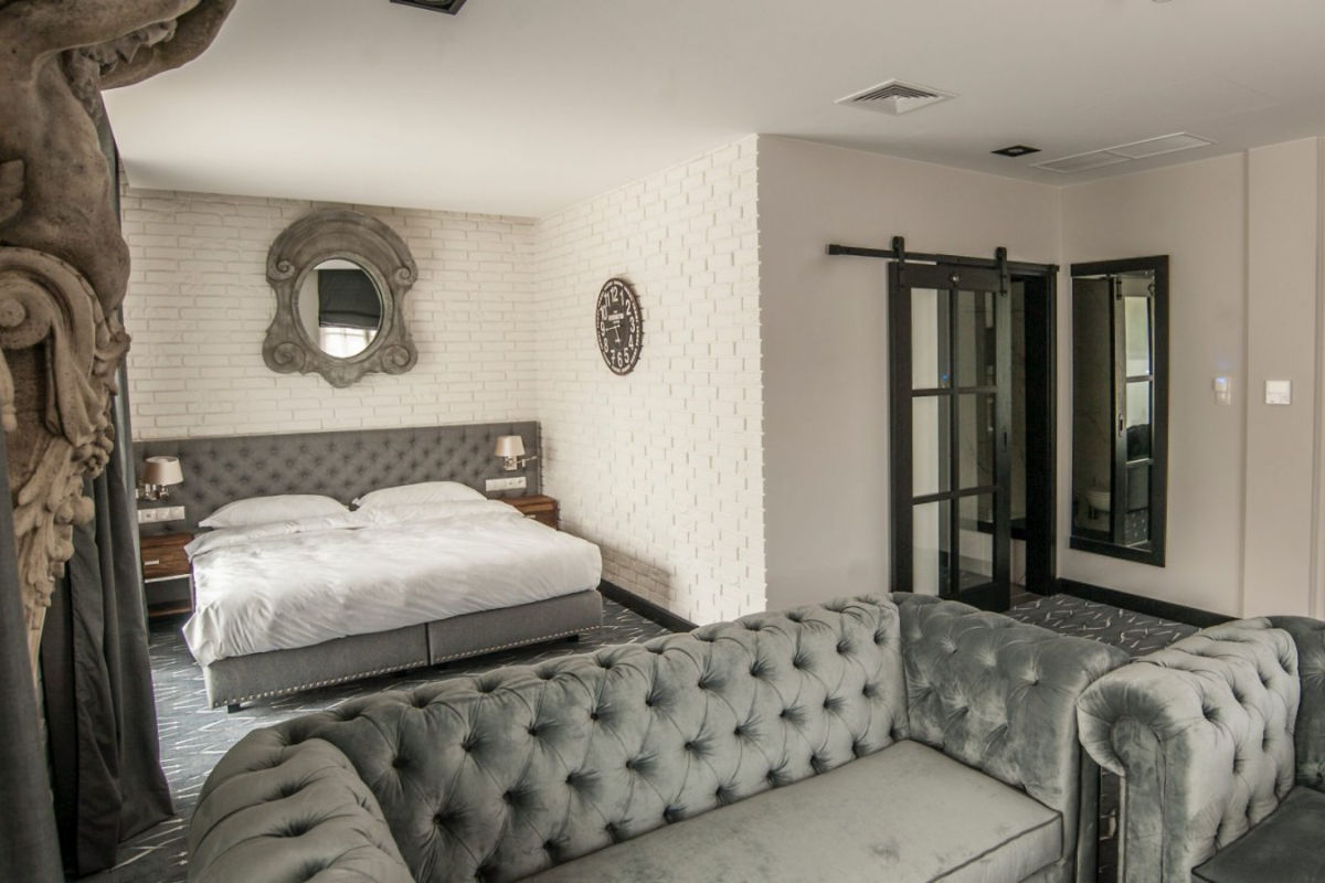 Una camera da letto moderna con un divano grigio in primo piano, un grande letto con biancheria bianca e uno specchio sopra la testata. Ci sono porte a sbarramento che rivelano un armadio o un'altra stanza.