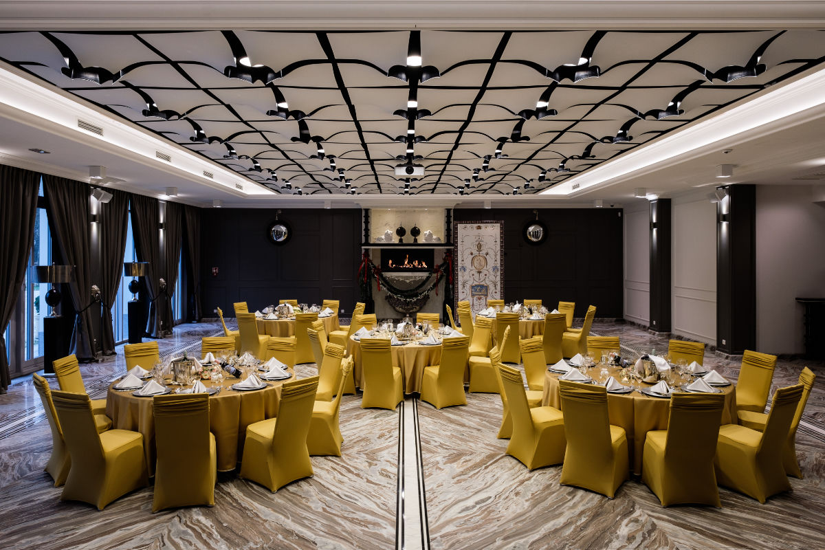 Una sala banchetti con sedie gialle e tavoli rotondi apparecchiati per la cena, con lampadari, finestre drappeggiate e un caminetto decorativo al centro.