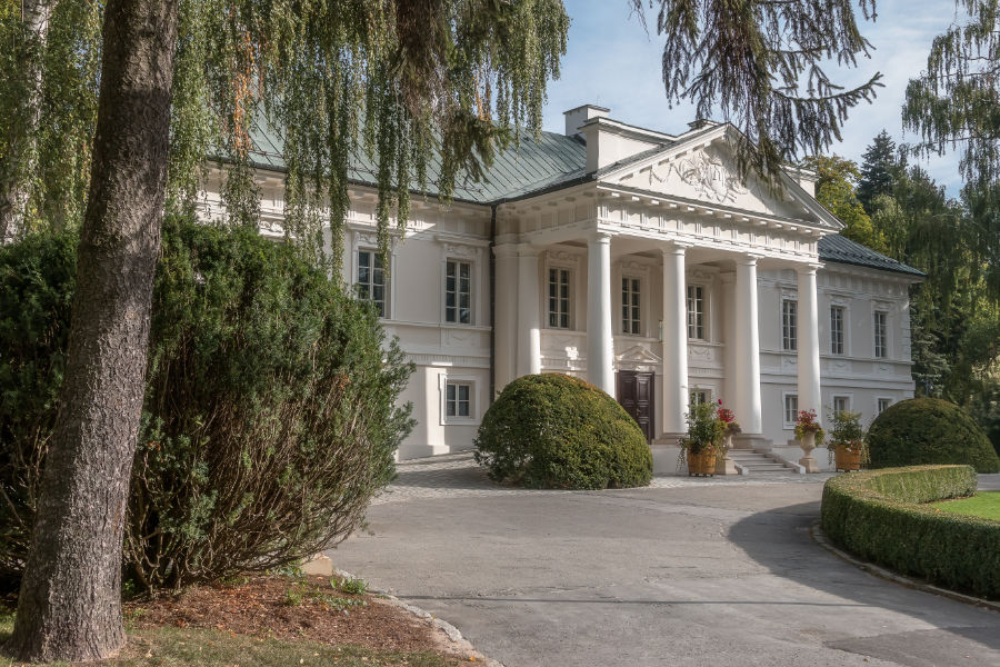 A large, white neoclassical mansion with four columns at the entrance, surrounded by well-manicured bushes and trees, reminiscent of Pałac Mała Wieś.