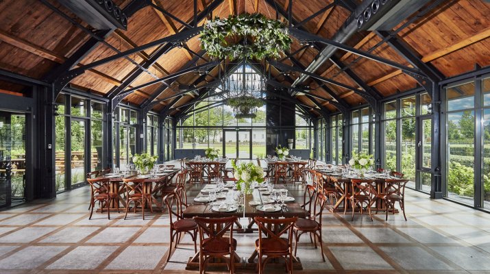 Una spaziosa e rustica sala per eventi del Pałac Mała Wieś con alti soffitti in legno, grandi finestre e tavoli apparecchiati con fogliame verde e fiori bianchi.