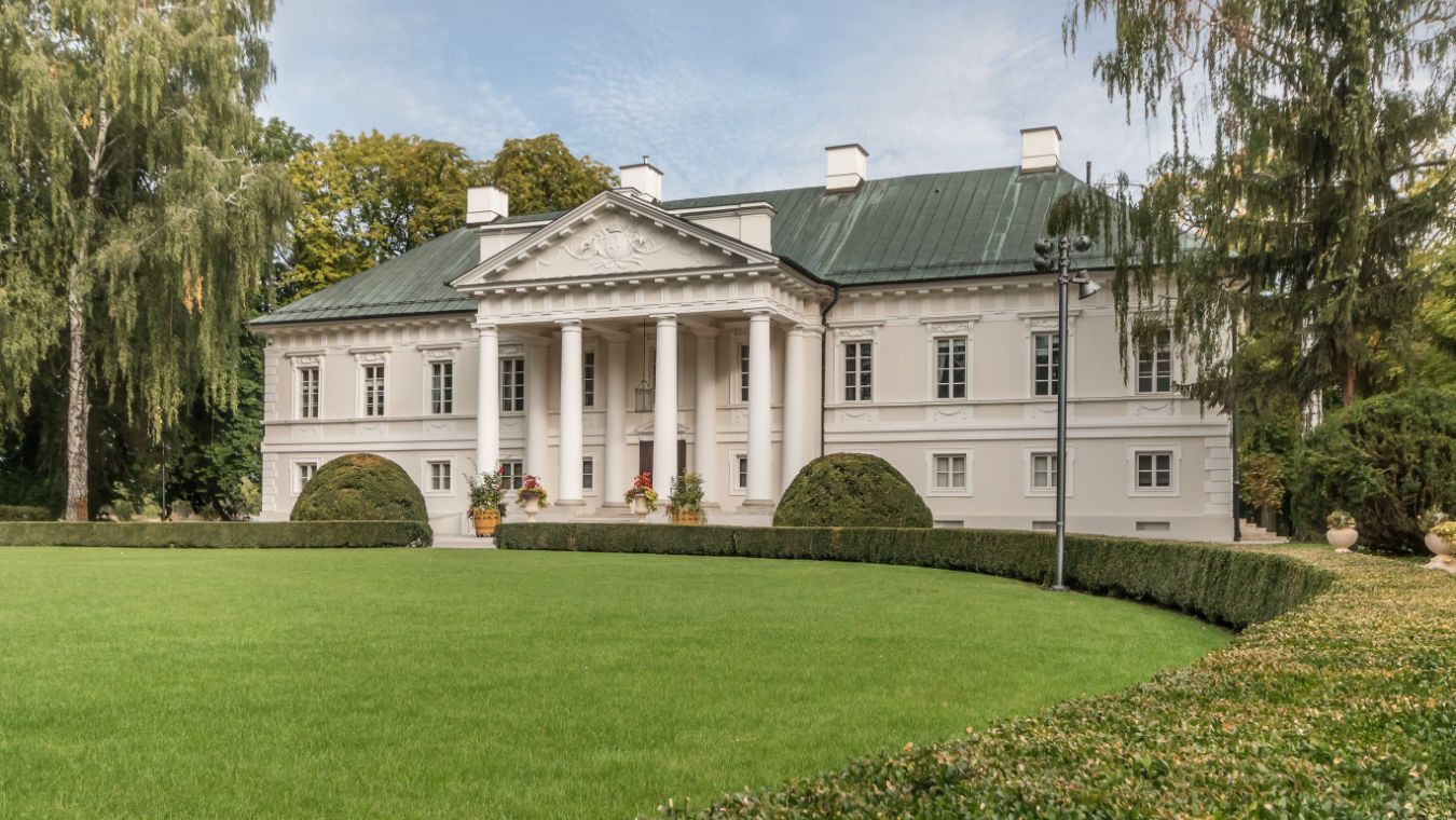 Una gran mansión neoclásica blanca con cuatro columnas, un tejado verde y un entorno ajardinado bajo un cielo azul.