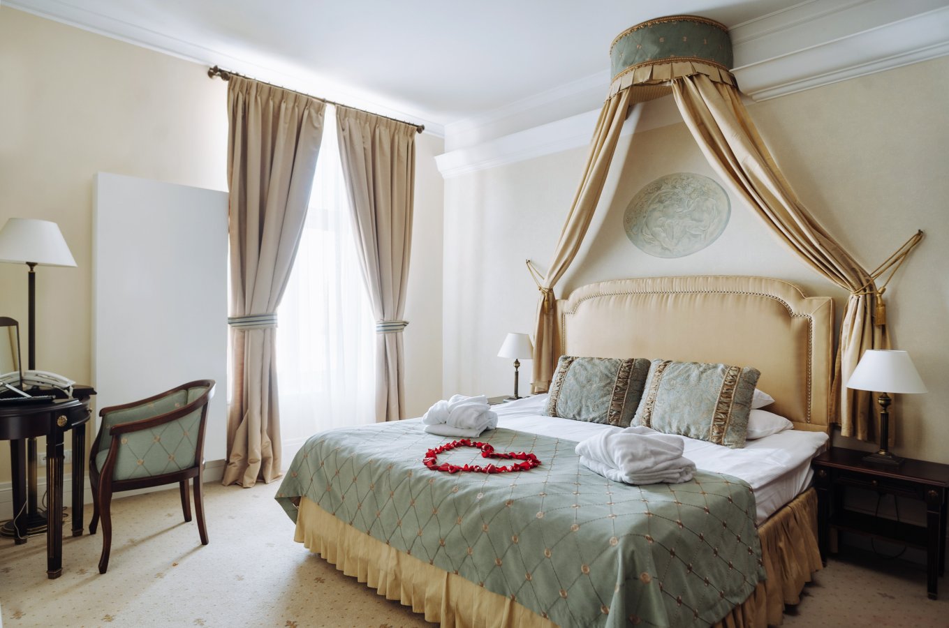 La lussuosa camera da letto dell'Hotel Pałac Romantyczny dispone di un letto king size ornato da asciugamani bianchi e da una decorazione a forma di cuore. La camera vanta un baldacchino, una scrivania con sedia e due comodini con lampade. La luce entra dalla finestra, creando un ambiente sereno.