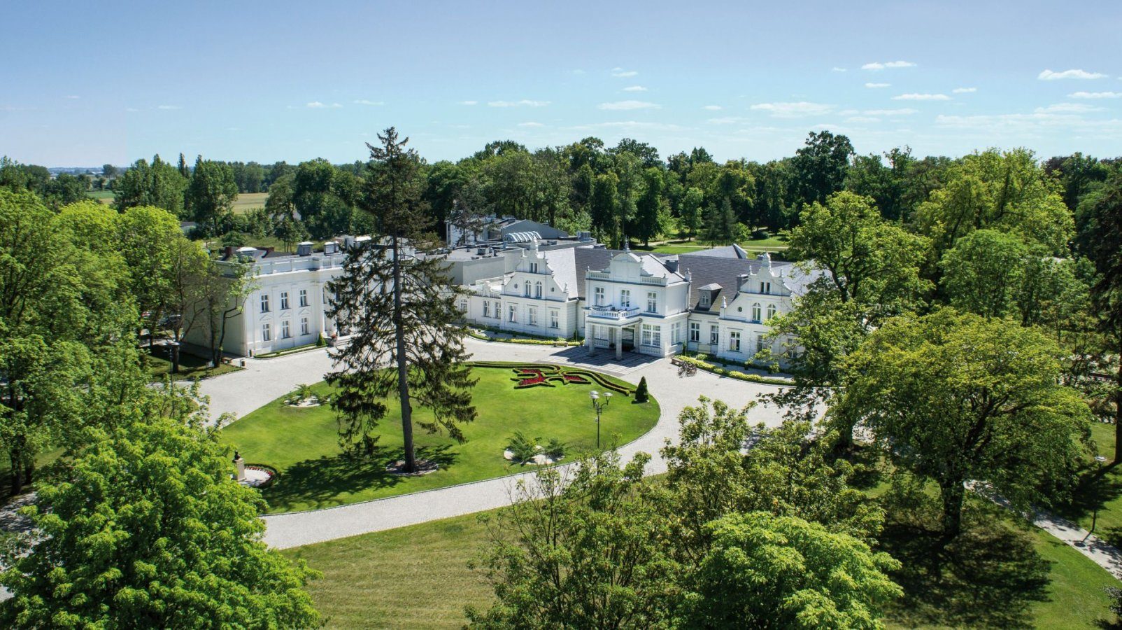 Veduta aerea dell'Hotel Pałac Romantyczny, un grande palazzo bianco con più ali, circondato da giardini, alberi e un viale circolare.