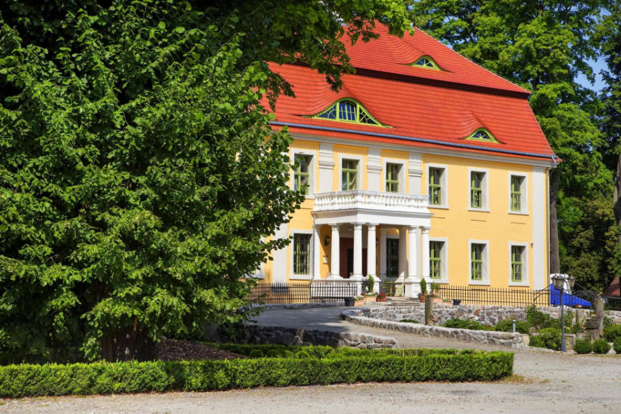 Le Pałac Wiechlice est un grand bâtiment jaune avec un toit rouge, des colonnes blanches à l'entrée et des cadres de fenêtres verts, entouré d'arbres et d'un chemin de gravier au premier plan.