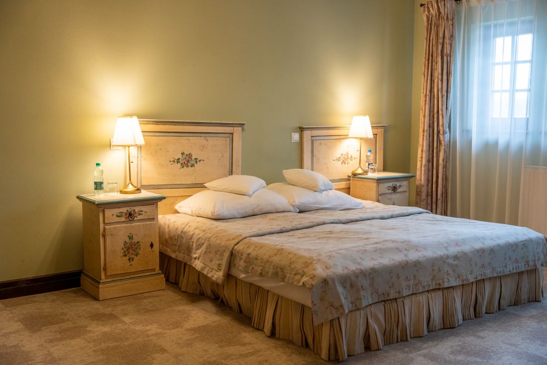 Ein Schlafzimmer mit einem Doppelbett, zwei Nachttischen mit Lampen und einem Blumendekor, das an Pałac Wojanów erinnert. Auf der rechten Seite befindet sich ein Fenster mit transparenten Vorhängen.
