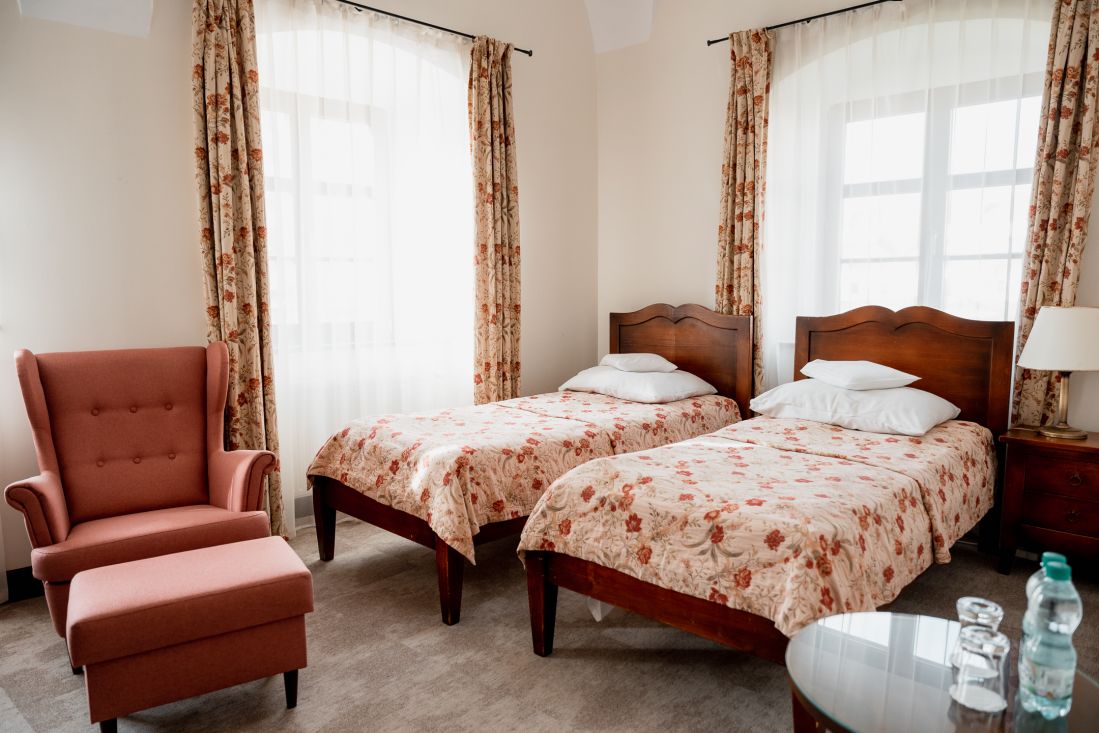 Ein Zweibettzimmer im Pałac Wojanów verfügt über zwei Einzelbetten aus Holz, geblümte Tagesdecken, einen pfirsichfarbenen Sessel mit Ottomane, geblümte Vorhänge, einen kleinen Tisch mit Wasserflaschen und eine Lampe auf einem hölzernen Nachttisch.