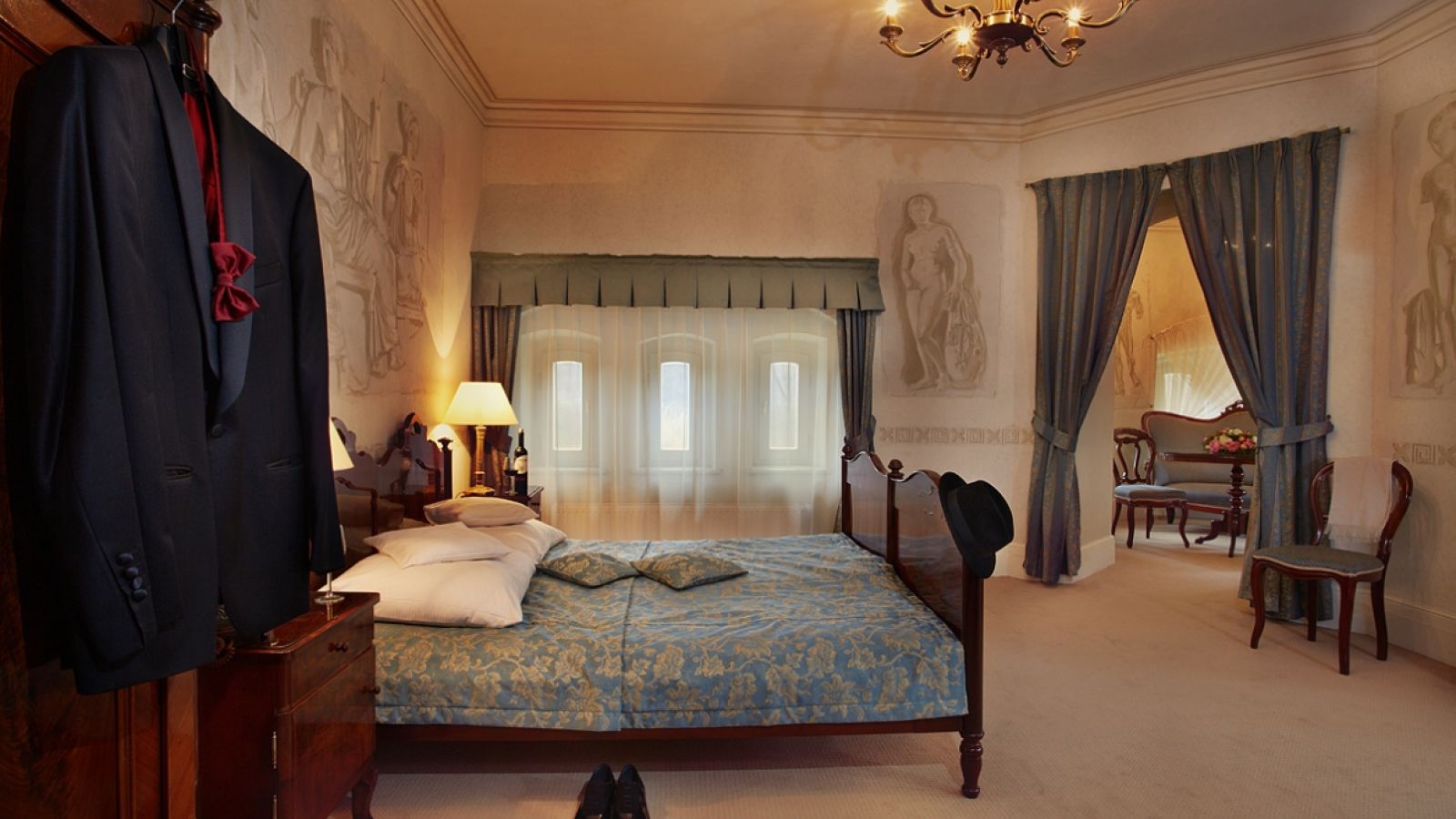 Ein helles Schlafzimmer in Pałac Wojanów mit einem Doppelbett, kunstvollem Dekor, einem Anzug im Kleiderschrank, Schuhen auf dem Boden und einer angrenzenden Sitzecke mit Tisch, Stühlen und Vorhängen.