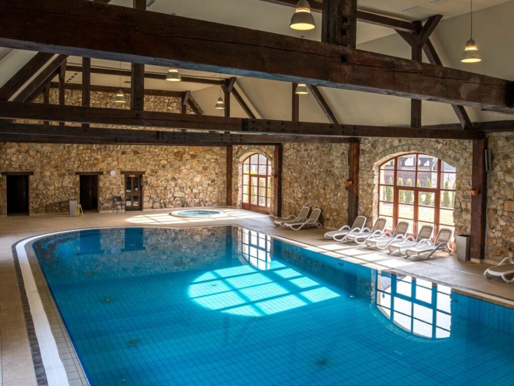 Fughe fresche: Dove alloggiare quando fa troppo caldo in città 3 Piscina coperta con pareti in pietra, travi in legno, pavimento piastrellato e sedie a sdraio che ricordano Pałac Wojanów. Grandi finestre ad arco permettono alla luce naturale di illuminare l'area della piscina.