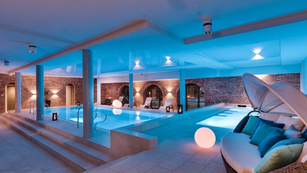 Wellness-Saison: Wärmende Rückzugsorte in historischen Hotels in Europa 12 Entdecken Sie den weitläufigen Innenpool des Talaria Resort & SPA mit moderner Beleuchtung, eleganten Backsteinwänden, luxuriösen Pool-Liegen und einem gemütlichen Sitzbereich mit Kissen und Baldachin.