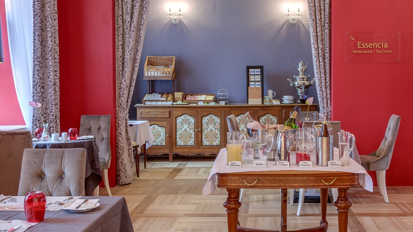 Ein Raum mit einem Frühstücksbuffet im Talaria Resort & SPA. Auf zwei Tischen stehen verschiedene Getränke und Speisen. An der Wand hängt ein Schild mit der Aufschrift "Essencia".