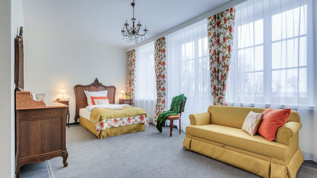 Wellness-Saison: Wärmende Rückzugsorte in historischen Hotels in Europa 11 Ein geräumiges Schlafzimmer im Talaria Resort & SPA ist mit einem Bett, einer Holzkommode, einem gelben Sofa und einem Stuhl ausgestattet. Das Zimmer verfügt über große Fenster, die mit bunten Blumenvorhängen geschmückt sind, und einen Kronleuchter, der von der Decke hängt.