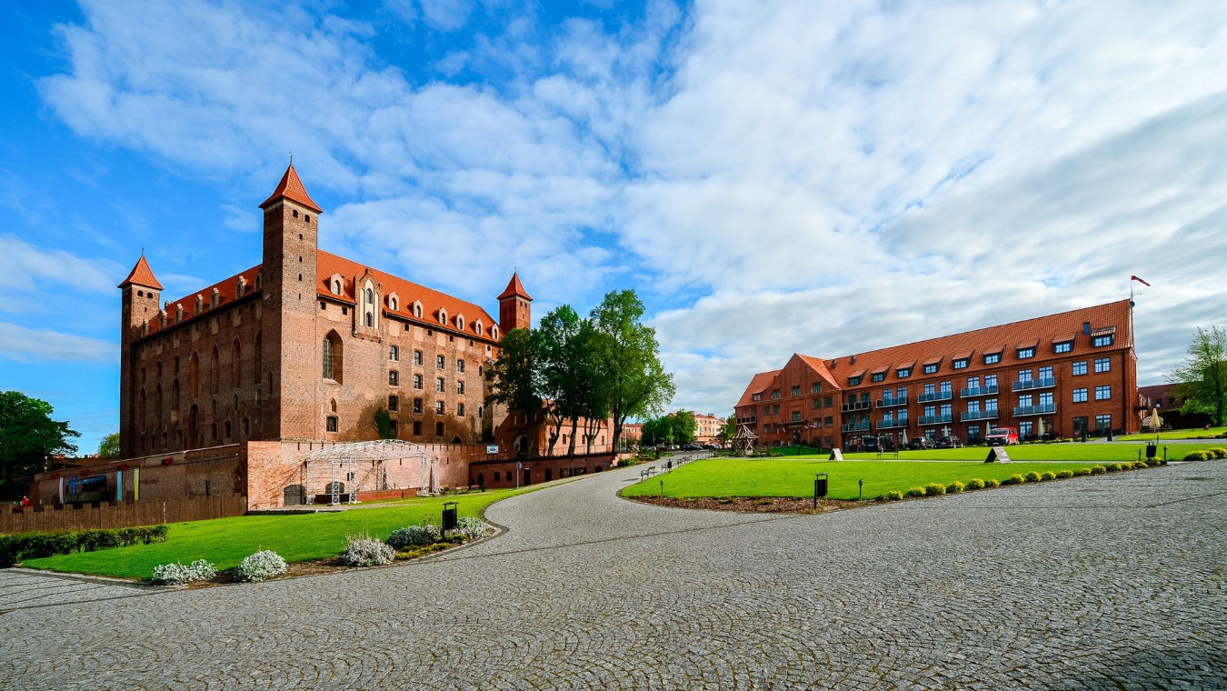 Un grand château historique au toit de tuiles rouges et aux multiples tours se dresse à côté du bâtiment moderne en briques de l'hôtel Zamek Gniew, tous deux sur fond de pelouses vertes et d'allées pavées, sous un ciel partiellement nuageux.