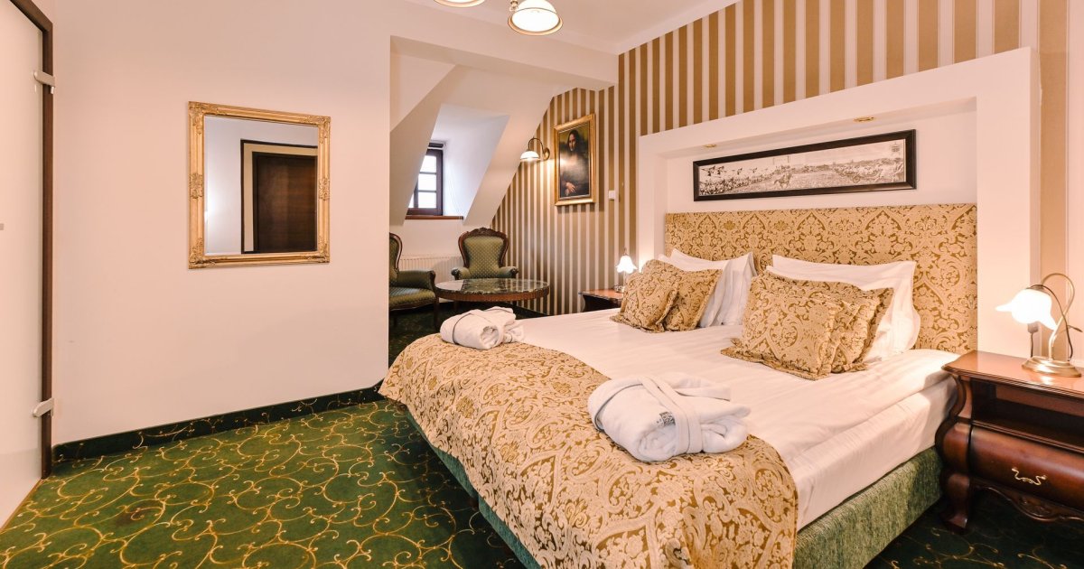 Camera d'albergo dell'Hotel Zamek Gniew con letto matrimoniale ornato da lenzuola verdi e dorate, due comodini con lampade e uno specchio a parete. L'accogliente area salotto comprende due sedie e un tavolo, il tutto sullo sfondo di un tappeto a motivi verdi.