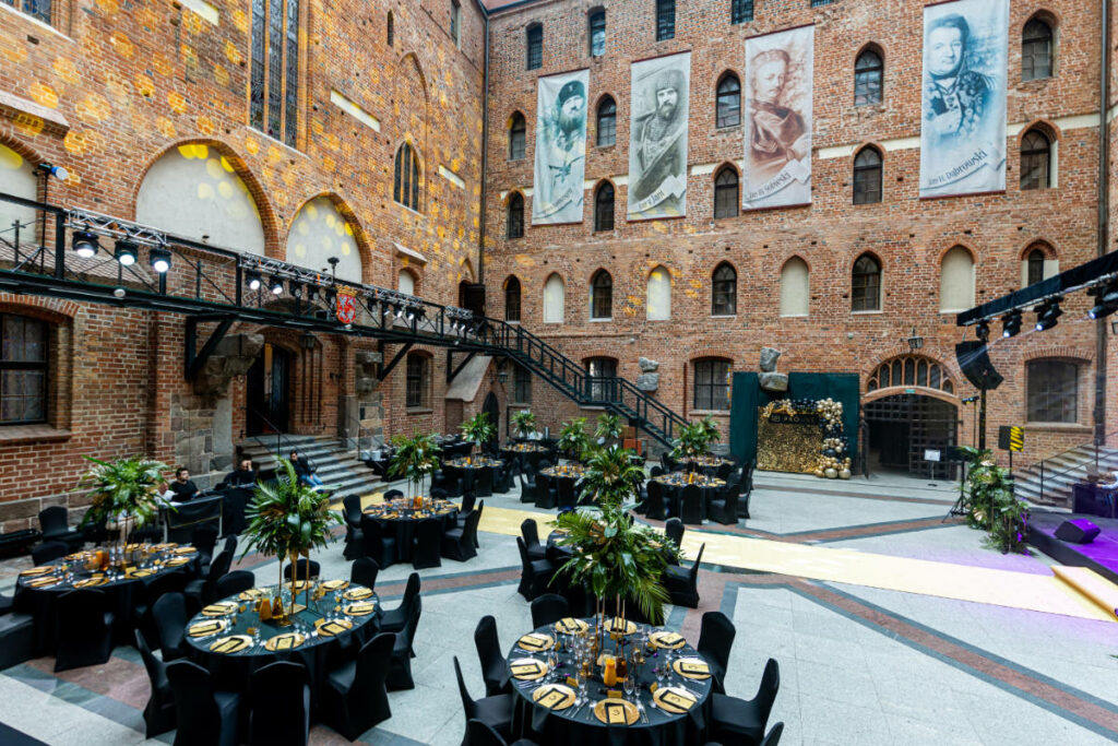 Europe’s Best-Kept Secrets: Lesser-Known Historic Hotel Escapes 20 A brick indoor venue at Hotel Zamek Gniew, arranged for an event with round tables set for dinner, decorated with flowers and candles. Portraits hang on the wall above a wooden staircase and a section of overhead lighting.