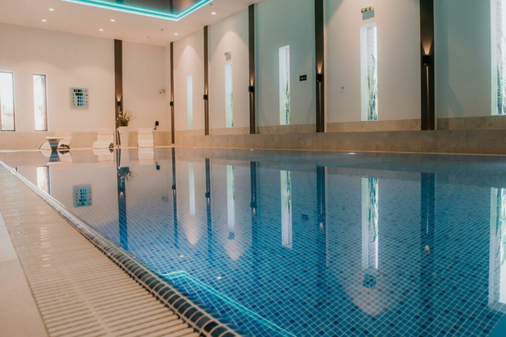 Europe’s Best-Kept Secrets: Lesser-Known Historic Hotel Escapes 21 Indoor swimming pool with blue tiles, modern lighting, and poolside seating area with white chairs and a small table at Hotel Zamek Gniew. The walls have narrow windows and emergency exit signs.