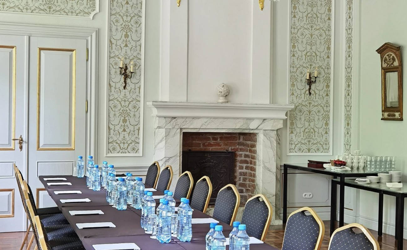 Salle de réunion formelle au sein du Zamek Janów Podlaski, dotée d'une longue table, de chaises, de bouteilles d'eau et de blocs-notes. L'élégance de la pièce est soulignée par une cheminée, des murs ornés, deux portes et une table supplémentaire avec des tasses et des assiettes.