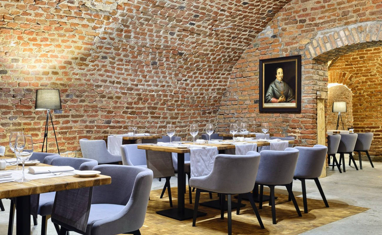 Ce restaurant aux murs de briques dispose de tables en bois garnies de linge blanc et de couverts bien rangés. Des chaises grises modernes et une photo d'un personnage historique du Zamek Janów Podlaski ornent la salle.