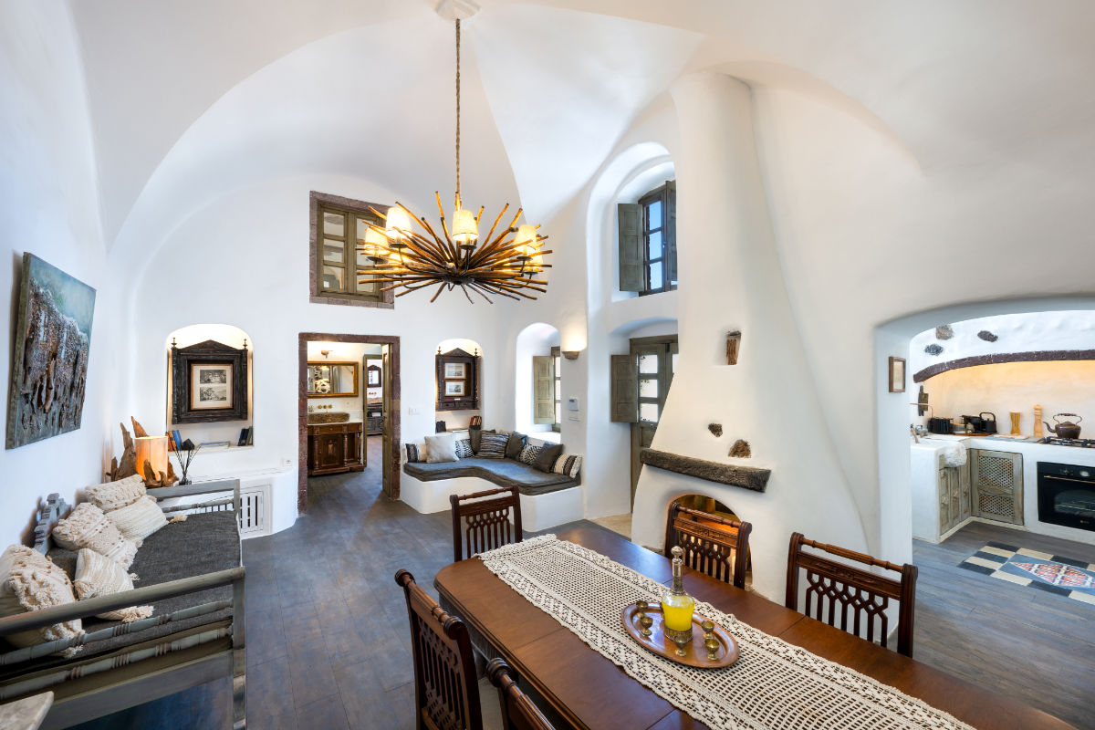 Spacious white room at Villa Santa Croce with an arched ceiling, modern chandelier, wooden dining table, cushioned benches, art pieces, and a kitchen area in the background.